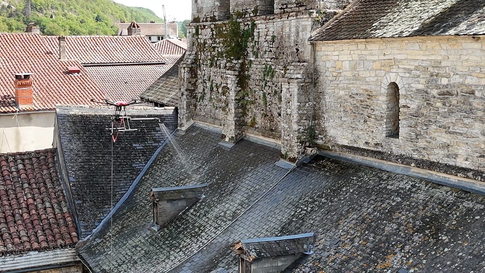 Vue aérienne d'ensemble : drone en plein traitement sur une toiture ancienne, village en arrière-plan
